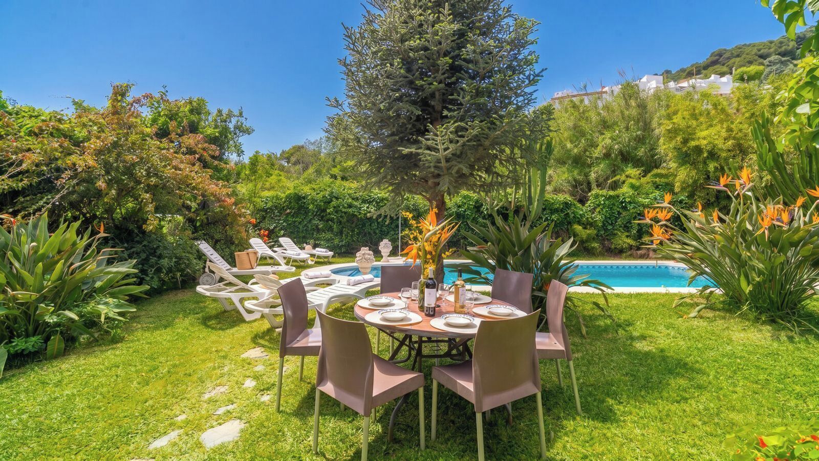 A poolside dining area with a table set for four, surrounded by lush greenery and a clear blue sky.