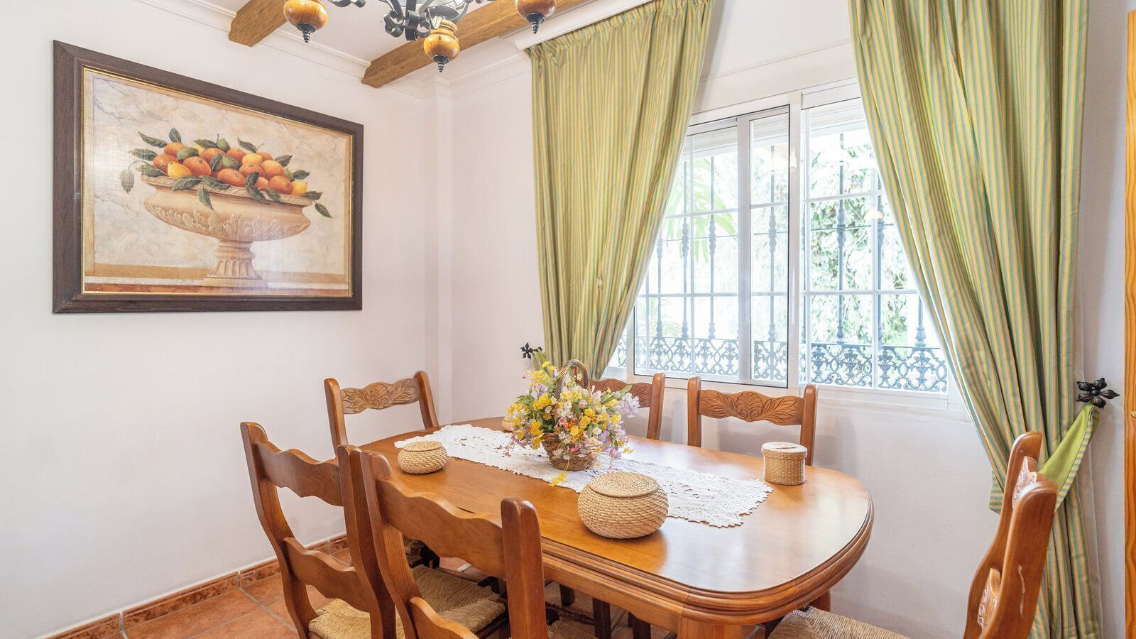 Une salle à manger avec une table en bois, des chaises, un vase de fleurs, un tableau encadré et des rideaux verts.