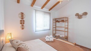 A bedroom with a bed, a wooden shelf with towels, a window with blinds, and wall decorations.