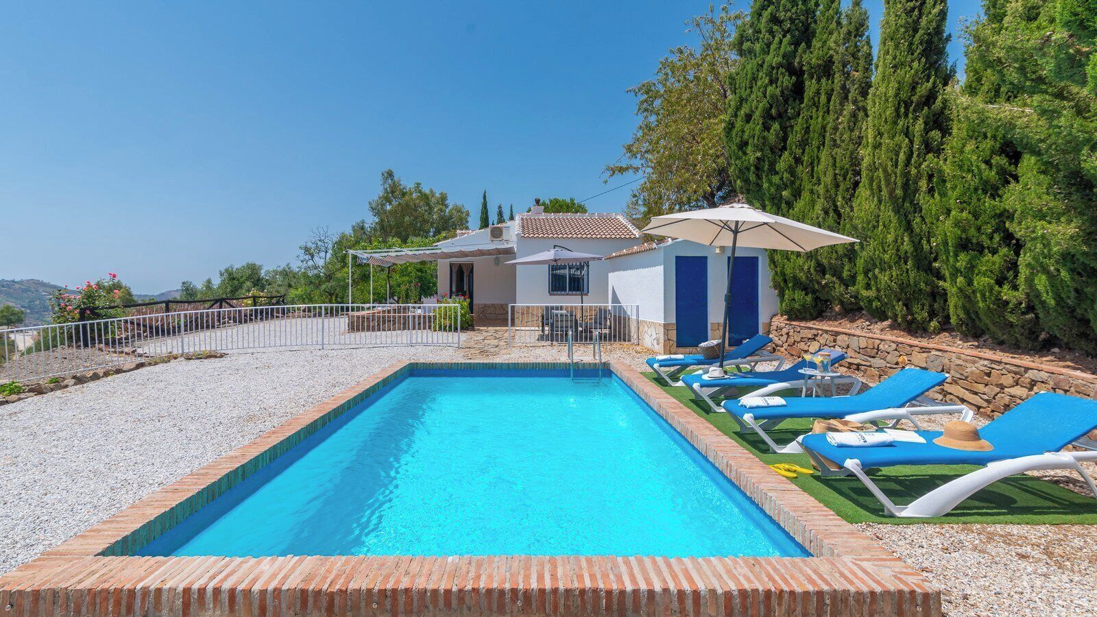 A rectangular swimming pool with a tiled edge, surrounded by a gravel area and a row of blue lounge chairs with umbrellas.