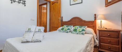 A neatly made bed with white linens and two towels with a logo, a wooden nightstand with a lamp, and a wooden door in the background.