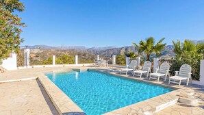 A rectangular pool with clear blue water, surrounded by white lounge chairs and palm trees, with a mountainous landscape in the background.
