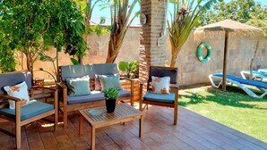 An outdoor patio area with wooden furniture, including a sofa and chairs, a small table, and a thatched-roof structure.