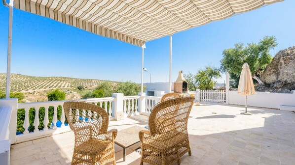 A covered outdoor seating area with wicker chairs and a table, overlooking a hilly landscape. - Almenara Los Corrales by Ruralidays (Seville)