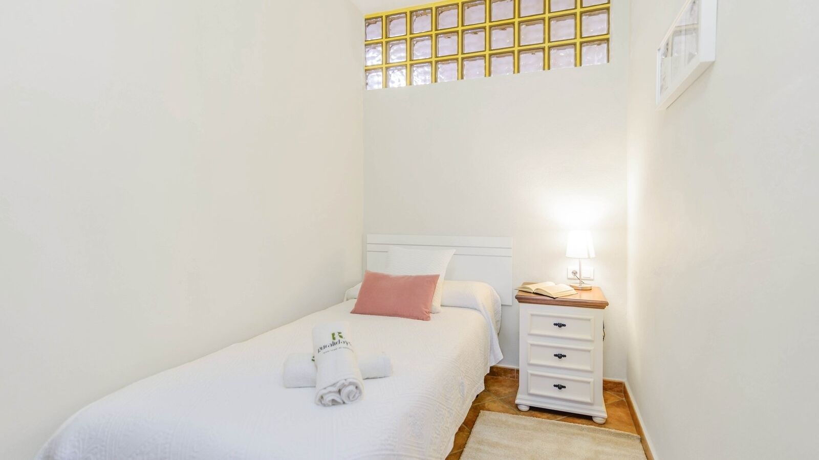 A small, clean room with a single bed, a nightstand with a lamp, and a window with a grid pattern.