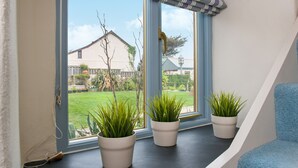 Una ventana con tres plantas en macetas sobre un alféizar, una silla azul y vista a una casa y jardín.