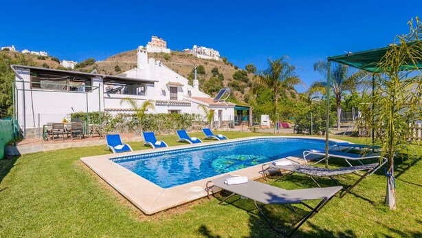 Una piscina amb gandules, una zona enjardinada i un edifici amb un turó al fons.