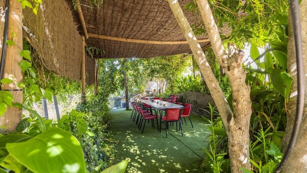 Een buiten eethoek met een tafel en stoelen, omgeven door weelderige begroeiing en een rieten dakconstructie.