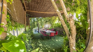 An outdoor dining area with a table and chairs, surrounded by lush greenery and a thatched roof structure.