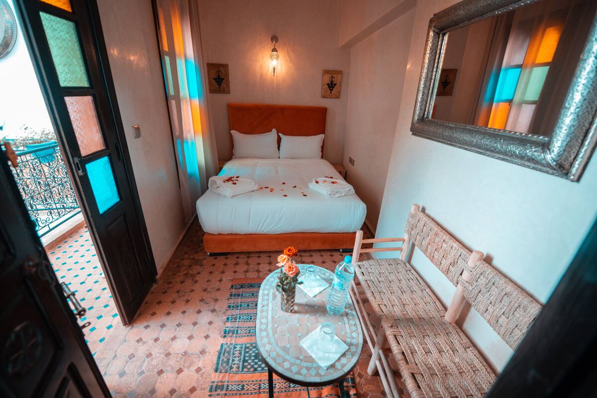 Habitación de hotel con cama, una pequeña mesa con un florero y una botella, una silla de mimbre y un balcón con barandilla.