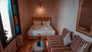 Une chambre d’hôtel avec un lit, un canapé, une petite table avec un vase et des bouteilles, et une fenêtre avec des rideaux.