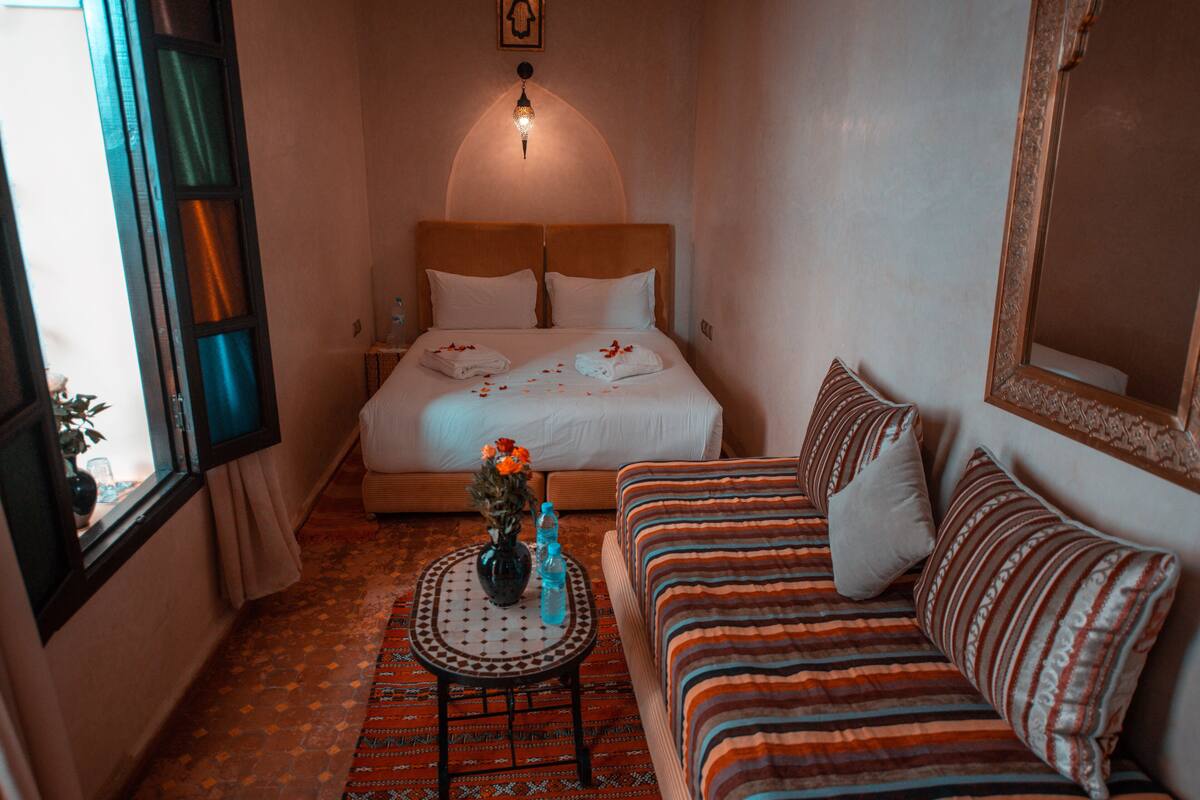 Habitación de hotel con cama, sofá, una mesita con un jarrón y botellas, y ventana con cortinas.