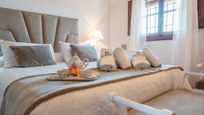 Quarto de hotel com uma cama grande, mesa de cabeceira com bule e chávenas, toalhas dobradas e janela com cortinas.