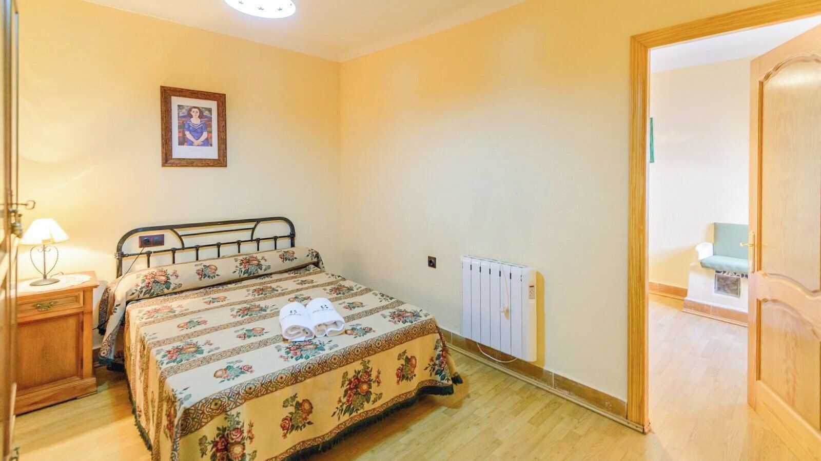 A bedroom with a bed, a radiator, a wooden door, and a small table with a lamp.