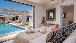 Un dormitorio con un ventanal grande que da a una piscina, una cama con sábanas blancas y un cojín estampado, y una mesita de noche con un espejo.