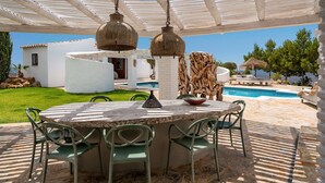 Un área de comedor al aire libre con una mesa redonda y sillas, un colgante de luz y una piscina al fondo.