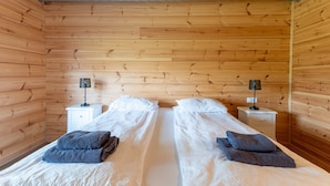 A room with wooden walls and two beds with white bedding and dark towels. There are two bedside tables with lamps. - Glaðheimar Guesthouse (Blönduós)