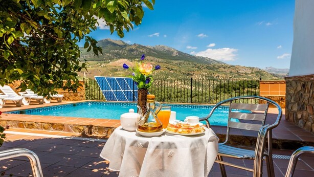 A poolside breakfast setup with a table, chairs, and a view of mountains.