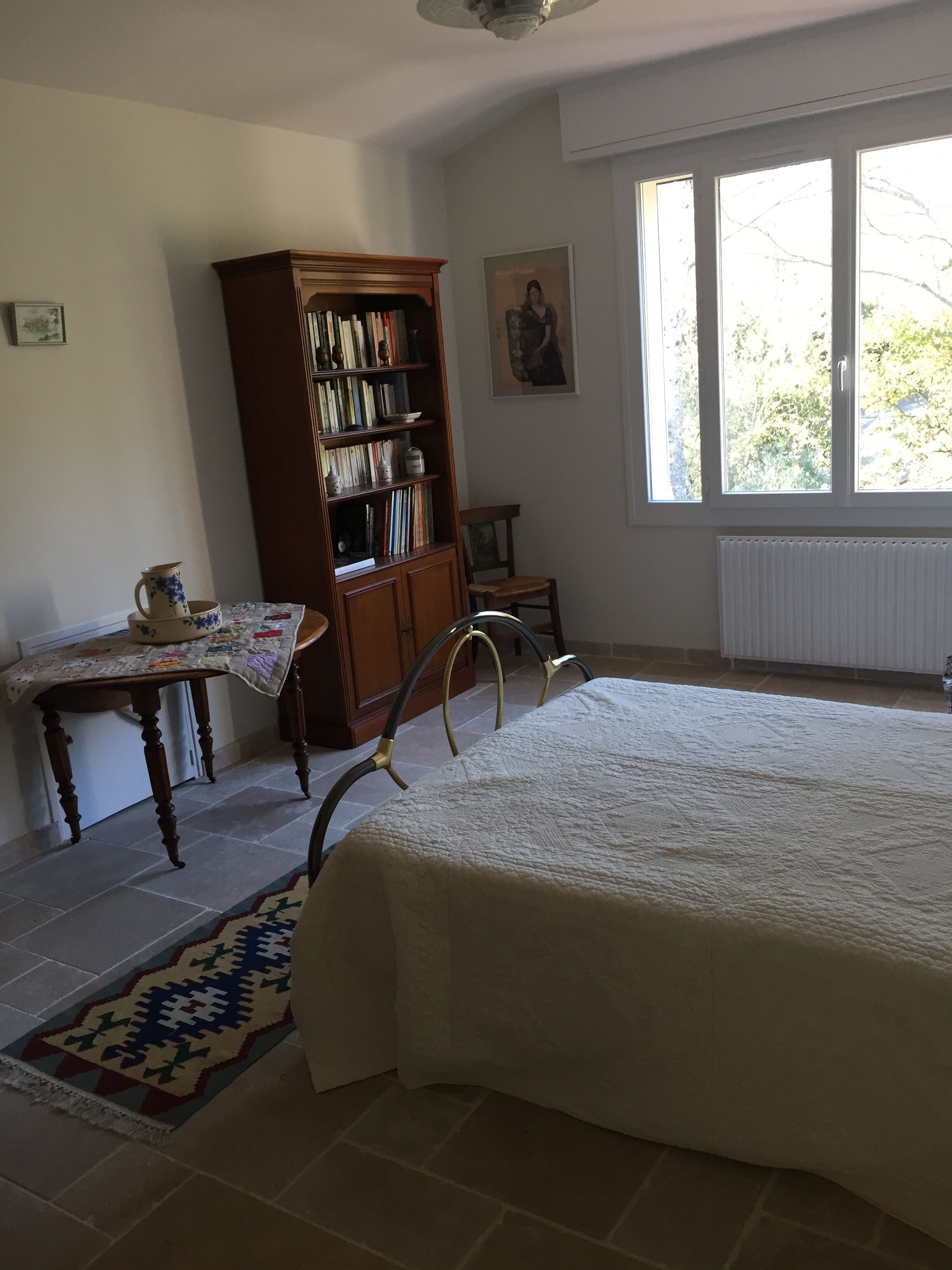 Un dormitorio con una cama grande, una estantería de madera, una mesita con una jarra, una silla y una ventana con vistas a árboles.