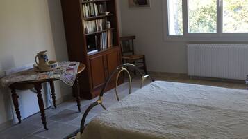 Un dormitorio con una cama grande, una estantería de madera, una mesita con una jarra, una silla y una ventana con vistas a árboles.