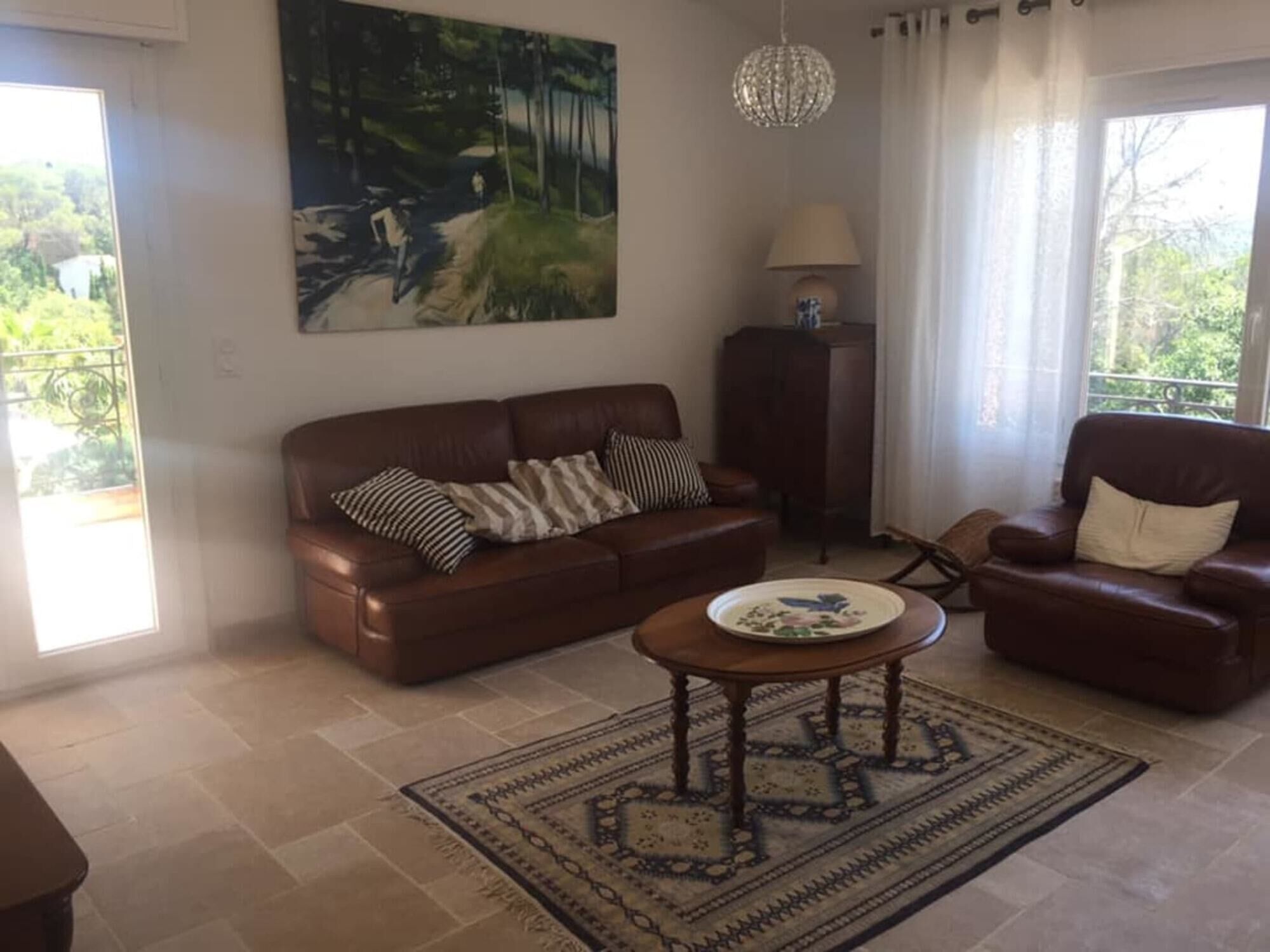 A living room with a brown leather sofa, a patterned rug, a table with a decorative plate, a painting of a forest scene, and a chandelier.