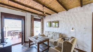 A cozy living room with a wooden table, sofa, and chairs, a balcony with outdoor seating, and a rustic ceiling.