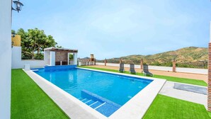 A rectangular swimming pool with a clear blue water, surrounded by artificial green grass and a white border. There are lounge chairs and a small structure with a tiled roof near the pool.