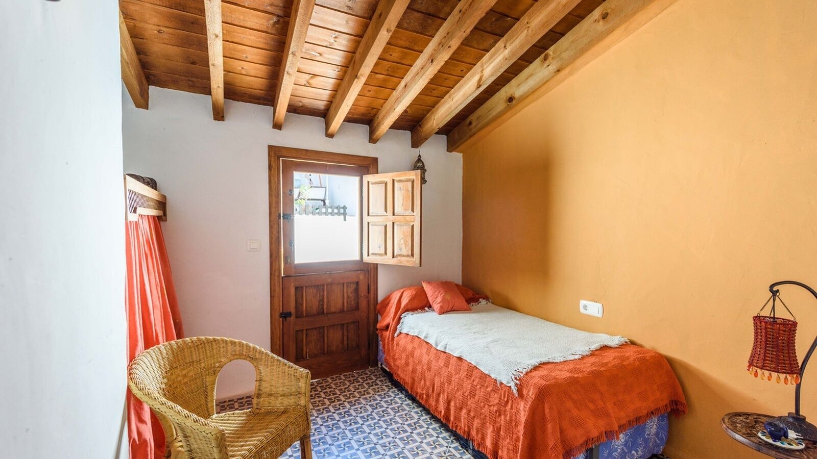 Ein gemütliches Schlafzimmer mit Holzboden, einem Bett mit orangefarbener Bettwäsche, einem Korbsessel und einem gemusterten Boden.