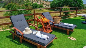 Two lounge chairs with cushions, a wicker basket, and a bottle of orange juice on a grassy deck with a wooden railing. - Huerta Alejo Alozaina by Ruralidays (Málaga)