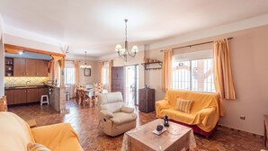 Un ampli espai de estar amb un sofà, una butaca i una taula de centre. Es veu una cuina amb armaris de fusta i una zona de dinar amb taula i cadires al fons.