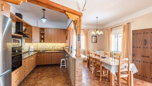 Cuina amb armaris de fusta, nevera, fogó i zona de dinar amb taula i cadires.