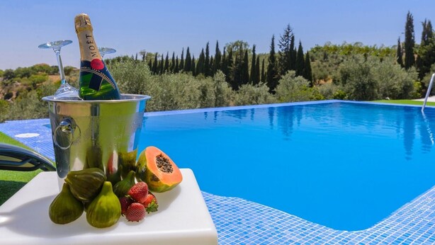Um ambiente junto à piscina, com uma garrafa de champanhe, frutas e vista desobstruída para a piscina e vegetação ao redor.