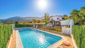 A swimming pool with a view of mountains, a fenced area with chairs, and a house in the background.