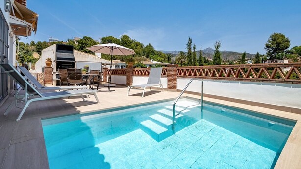 Ein Schwimmbecken mit Metallleiter, Liegestühlen und einer Terrasse mit Pergola und Sonnenschirm.