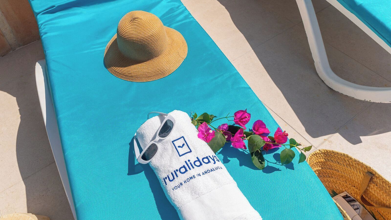 A poolside area with a blue mat, a straw hat, sunglasses, a towel with a logo, and pink flowers.