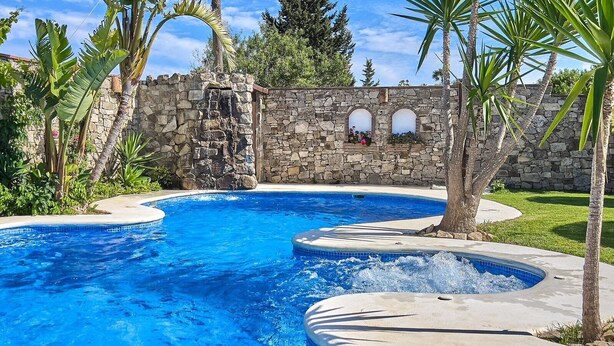 Une piscine bordée d’un mur en pierre et dotée de fenêtres en arc de cercle, entourée de plantes tropicales.