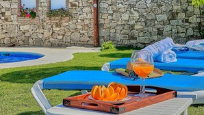 Un espace aménagé au bord de la piscine, avec des chaises longues, un plateau contenant des tranches d’orange et une boisson, et un mur de pierre en arrière-plan.