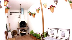 A cozy outdoor patio with a wooden bench, a small table, hanging planters with flowers, a rustic wood-fired oven, and a hanging lantern.