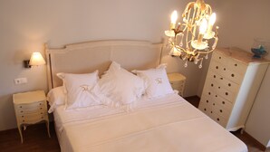 A neatly made bed with white linens and pillows, a chandelier above, a nightstand with a lamp, and a dresser with drawers.