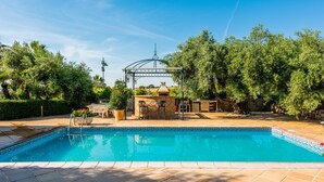 Ein Schwimmbad mit einer Pergola und einer Sitzgelegenheit im Freien, umgeben von Bäumen.