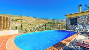 A swimming pool with a stone wall and a wooden deck, surrounded by a fence and chairs, with a view of hills in the background. - El Secano de Paco Órgiva by Ruralidays (Granada)