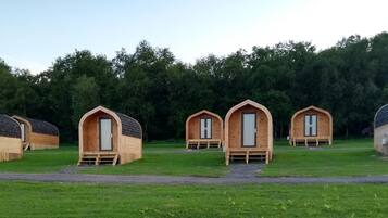 A row of wooden cabins with pitched roofs, arranged in a line on a grassy field with a forest in the background.