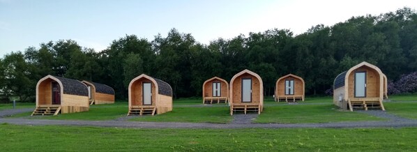 A row of wooden cabins with pitched roofs, arranged in a line on a grassy field with a forest in the background.