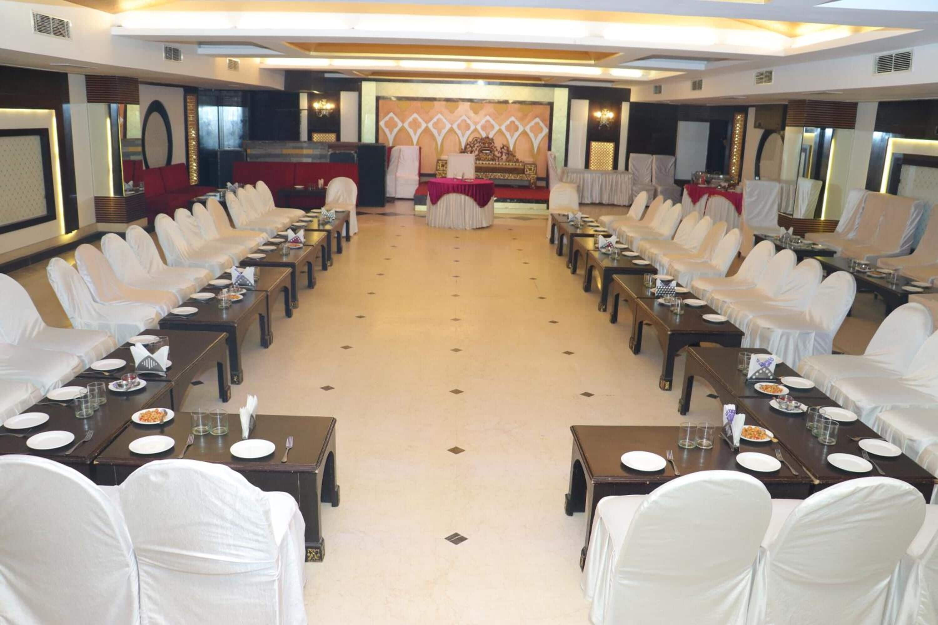 Un salón de banquetes con filas de sillas y mesas cubiertas de blanco, un escenario con un telón de fondo rojo y un espejo grande en la pared derecha.