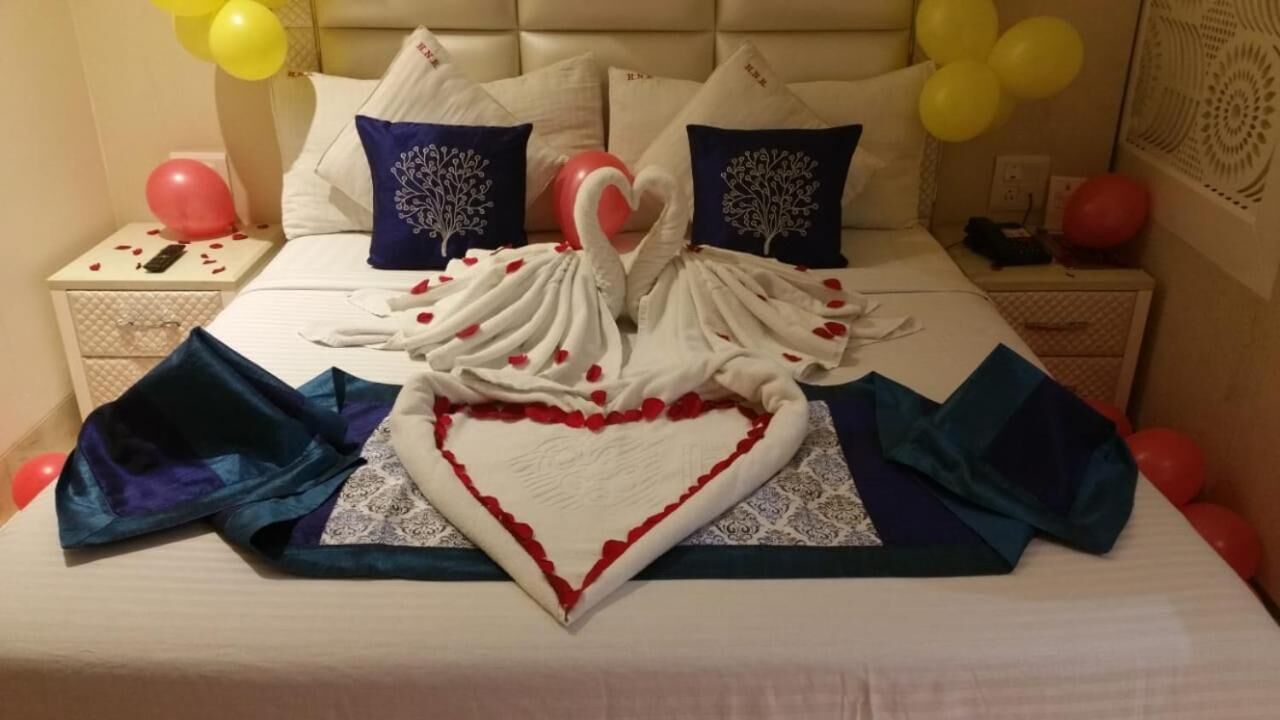 Una cama con un centro decorativo en forma de corazón hecho de tela y pétalos de rosa, rodeada de almohadas azules y blancas, y globos amarillos al fondo.
