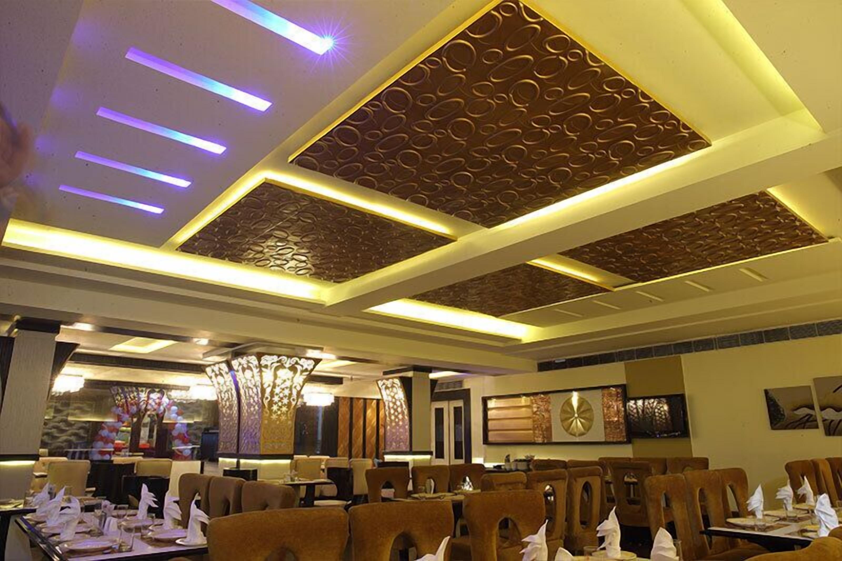 Restaurante con un diseño de techo moderno que incluye iluminación empotrada y paneles decorativos. Las mesas están dispuestas con servilletas y platos blancos.