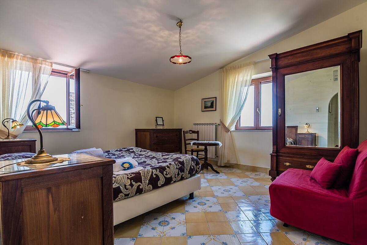 A bedroom with a bed, a wooden dresser, a red sofa, a mirror, and a lamp.