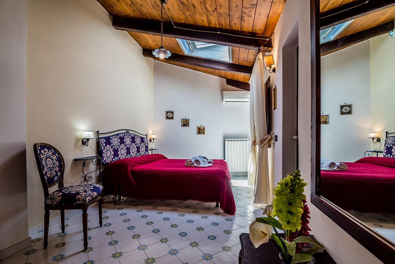 A bedroom with a wooden ceiling, a bed with a red bedspread, a chair with a patterned cushion, a small table with a vase of flowers, and a mirror reflecting the room.