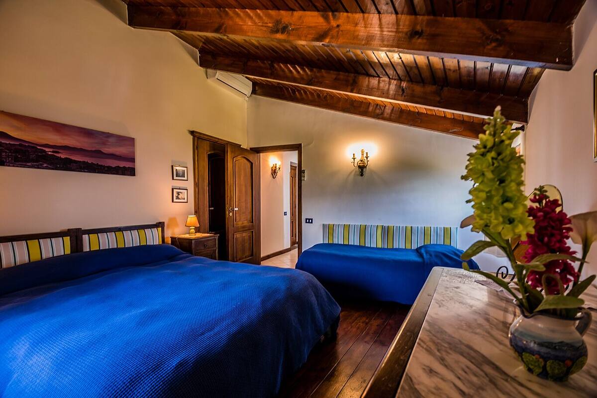 A hotel room with two beds, a wooden ceiling, a painting on the wall, and a vase with flowers on the table.