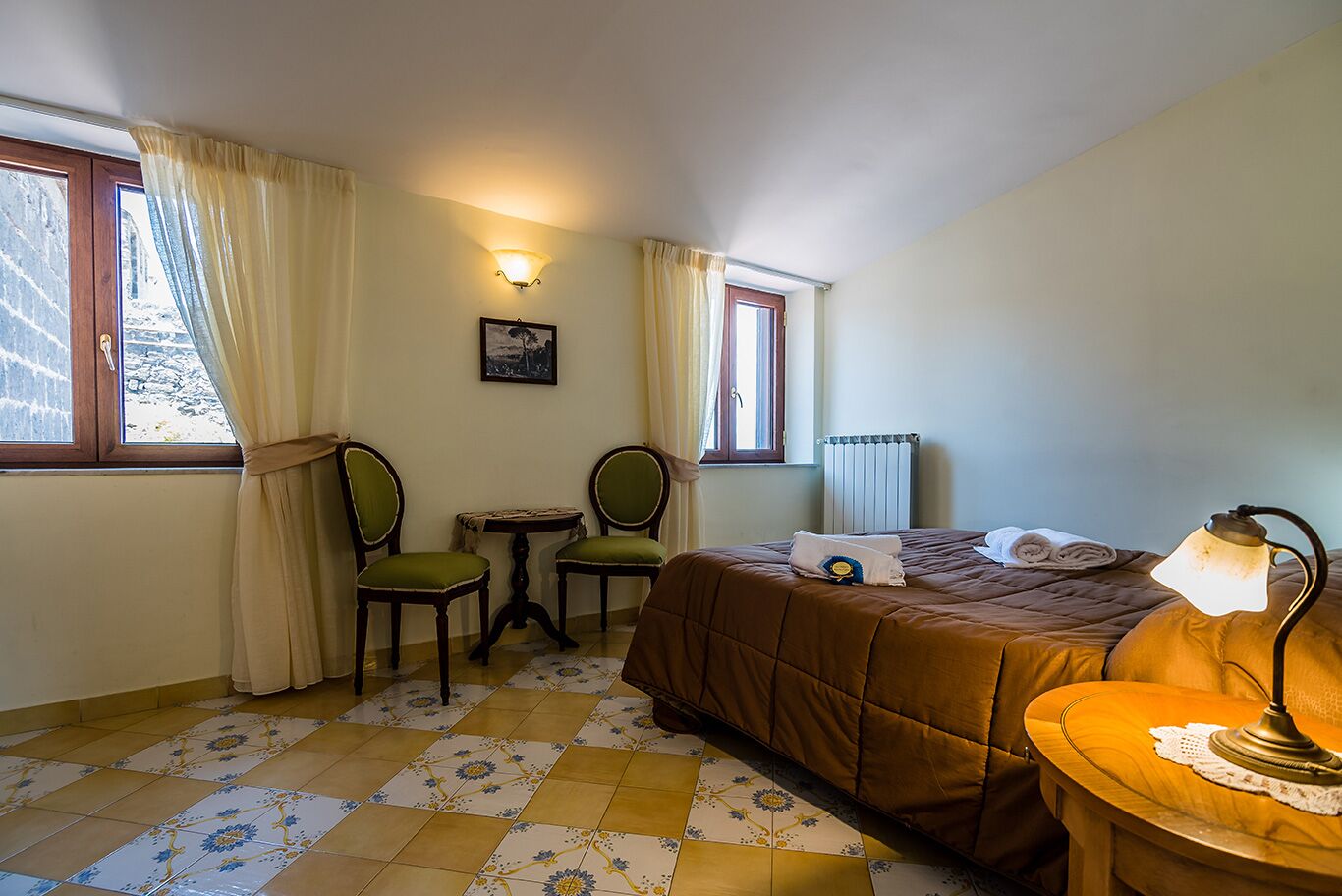 A hotel room with a bed, a small table with chairs, a radiator, and a window with curtains.